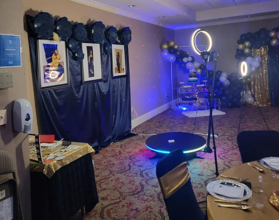 Indoor banquet hall party setup with navy-and-gold photo booth backdrop and framed portraits, black and gold balloon garlands, ring lights and LED rotating photo platform, cake table in the corner, and round dining tables with gold-accented chairs.