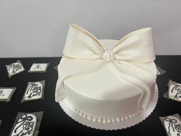 Elegant white fondant round wedding cake topped with an oversized silky bow and pearl bead border, displayed on a black table with individually wrapped bridal shower cookies.
