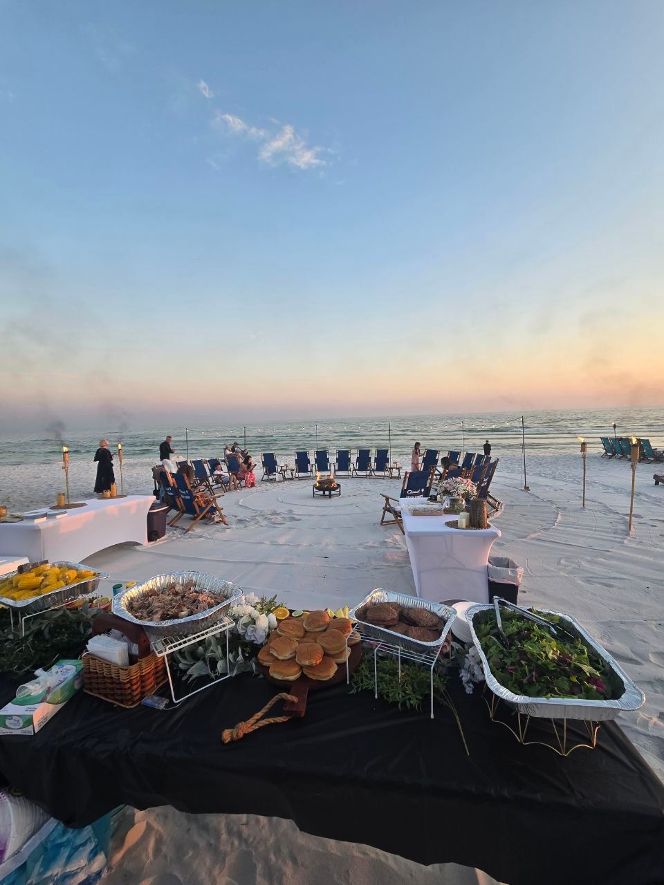 Oceanfront beach at sunset with a circle of folding chairs around a small bonfire, tiki torches lining the shoreline, and a foreground buffet table with buns, salad and sides set up for a casual seaside dinner.