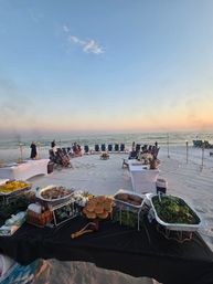 Oceanfront beach at sunset with a circle of folding chairs around a small bonfire, tiki torches lining the shoreline, and a foreground buffet table with buns, salad and sides set up for a casual seaside dinner.