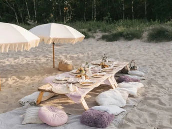 Boho beach picnic on sandy dunes with a low wooden table, pastel cushions and rugs, fringed parasols, place settings and small floral centerpieces by coastal grasses.