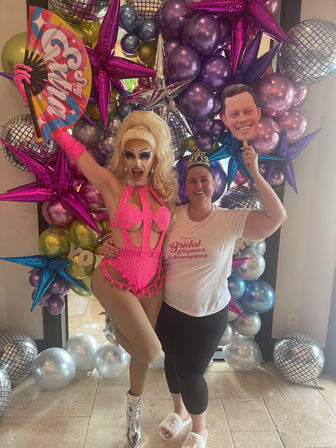 Glittering drag performer in a hot-pink rhinestone costume and towering blonde wig poses with a smiling guest wearing a tiara and holding a face cutout, colorful fan and silver boots in front of a metallic purple, blue and silver star-and-disco-ball balloon backdrop at a celebratory event.