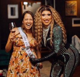 Two people laughing at a bachelorette party: a woman in a floral dress with a sash holding a wine glass and a smiling drag performer in a glittering bodysuit and long wavy hair posing indoors.