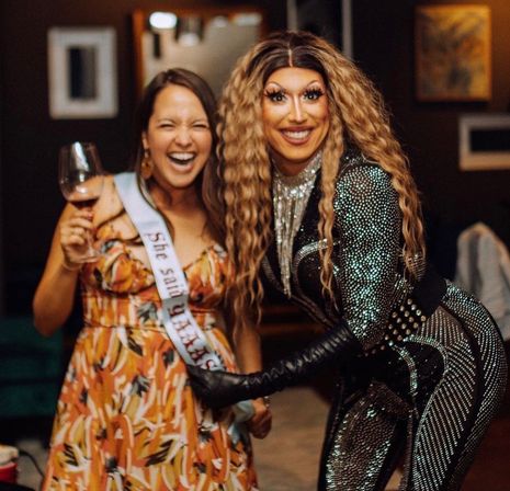 Two people laughing at a bachelorette party: a woman in a floral dress with a sash holding a wine glass and a smiling drag performer in a glittering bodysuit and long wavy hair posing indoors.