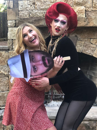 Drag performer in a sculpted red wig and black velvet outfit with gold chains poses playfully with a smiling blonde in a red floral dress holding a large cutout face in front of an outdoor stone fireplace.