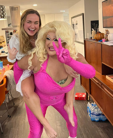 Drag performer in a bright pink jumpsuit and blonde wig flashes a peace sign while giving a laughing friend a piggyback ride at a cozy home kitchen party.