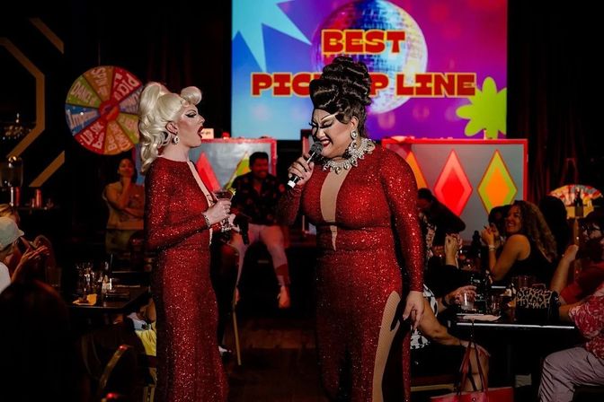 Two drag performers in sparkling red gowns onstage at a lively cabaret show, one singing into a microphone while the other holds a drink, colorful backdrop reads “BEST PICK UP LINE” and a seated audience watches and applauds.