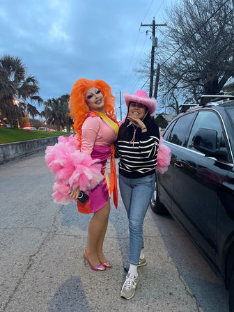 Drag performer in vibrant orange wig and pink ruffled dress poses smiling with a friend in a pink cowboy hat beside a black SUV on a palm-lined street at dusk.