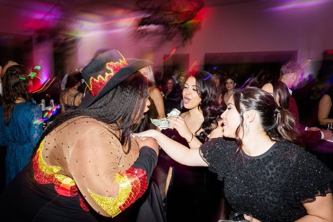 Lively indoor party scene: three women exchanging cash with a costumed performer wearing a flame‑decorated cowboy hat, colorful club lights and a crowded dance floor in the background.