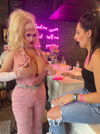 Drag performer in a pink star-studded jumpsuit and big blonde wig playfully covering their chest, surprising a seated woman at a neon-lit bar with a pink sign reading 'feed me tacos and tell me I'm pretty' — lively drag brunch vibe.