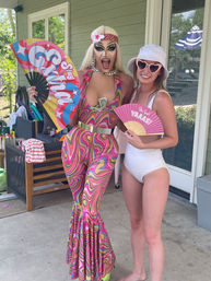 Drag performer in a colorful psychedelic 70s-style jumpsuit and blonde wig holding a bright fan, posing with a woman in a white swimsuit and heart-shaped sunglasses on a backyard poolside patio.