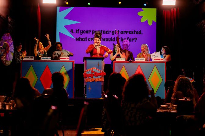 Drag queen host in a red sequined gown leads a lively game-show night on a colorful cabaret stage with contestants at numbered podiums cheering under bright stage lights and audience silhouettes
