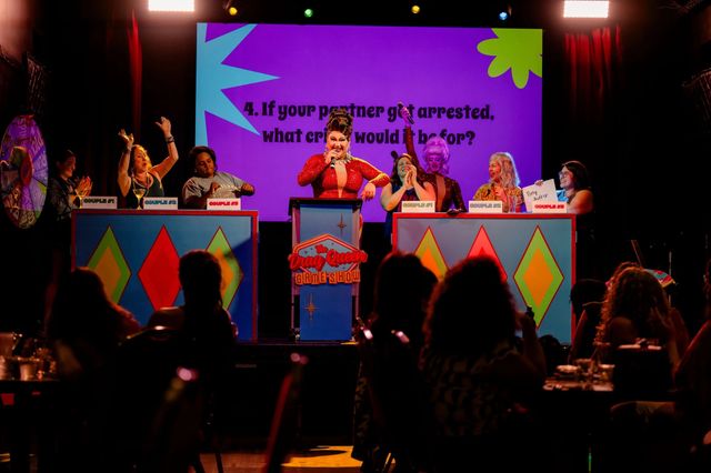 Drag queen host in a red sequined gown leads a lively game-show night on a colorful cabaret stage with contestants at numbered podiums cheering under bright stage lights and audience silhouettes