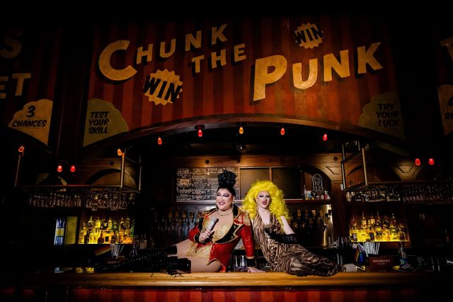 Two drag performers in glittering costumes pose on a wooden bar inside a dimly lit cabaret-style bar, with a retro carnival sign above, warm hanging bulbs, backlit liquor shelves and rows of hanging glasses.