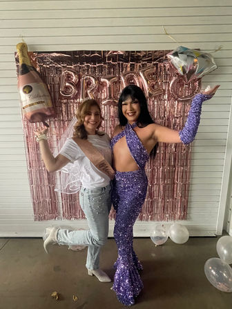 Two people smiling at a bachelorette party photo wall — one in a white tee, veil and rose-gold sash holding a champagne balloon, the other in a purple sequined cutout jumpsuit and matching glove, posing in front of rose-gold fringe and 'BRIDE' balloons with clear balloons on the floor.
