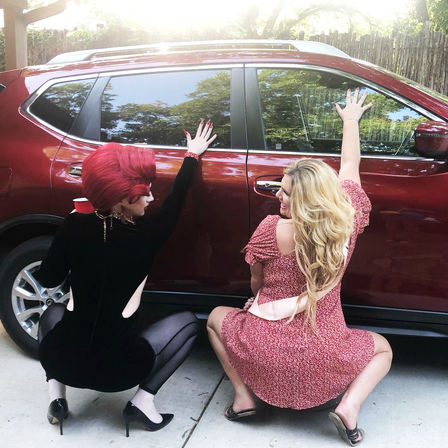 Two people playfully squatting and pressing hands against a shiny red SUV in a sunny driveway — one in a black outfit with a red wig and cup, the other in a pink floral dress with long blonde hair.