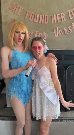 Bachelorette party: drag performer in a blue fringe costume and blonde wig poses with a bride-to-be in a white lace dress, pink sunglasses and sash, sharing a rhinestone microphone beneath a pink glitter “She Found Her…” banner.