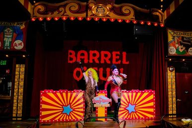 Two drag performers in glittery costumes and bold wigs on a colorful carnival-style cabaret stage with 'BARREL O FUN' marquee, starburst panels, retro marquee lights and a live-audience setup.