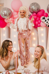 Drag performer in sparkly pink star pants and white ruffled top entertaining smiling guests at a pink balloon and disco-ball party celebration.