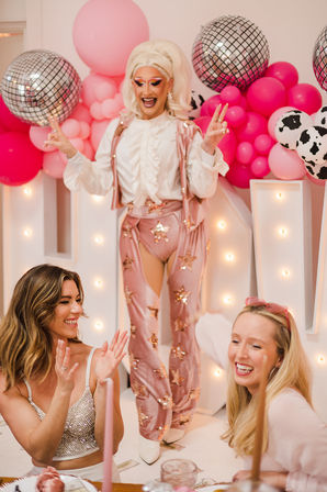 Drag performer in sparkly pink star pants and white ruffled top entertaining smiling guests at a pink balloon and disco-ball party celebration.