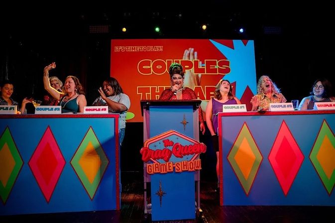Glittering drag performer in a red gown hosting a lively couples-themed game show on a colorful cabaret stage, with cheering contestants seated behind bright diamond-patterned podiums.