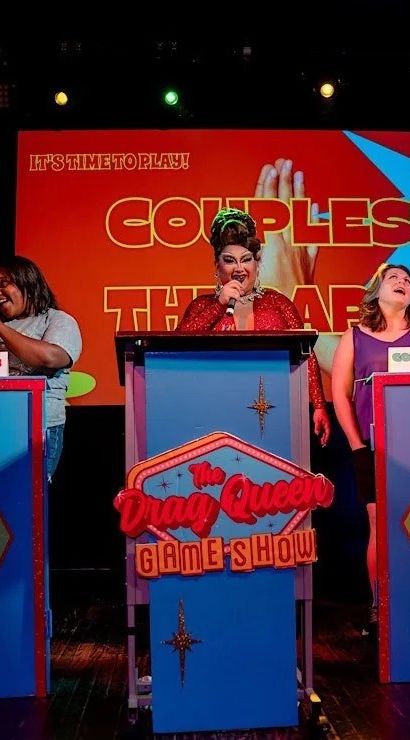Glittering drag performer in a red gown hosting a lively couples-themed game show on a colorful cabaret stage, with cheering contestants seated behind bright diamond-patterned podiums.