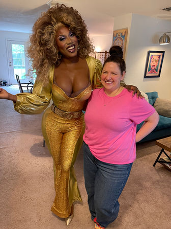 Two smiling people in a cozy living room — a glamorous performer in a gold sequined jumpsuit with voluminous curly hair posing beside a casually dressed friend in a pink T‑shirt and jeans.