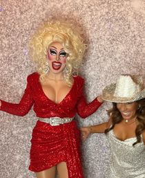 Glamorous drag performer in a sparkling red dress, big blonde wig and bold stage makeup laughing beside a woman in a silver sequin dress and glitter cowboy hat against a textured photo‑booth backdrop