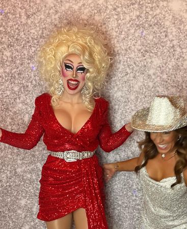 Glamorous drag performer in a sparkling red dress, big blonde wig and bold stage makeup laughing beside a woman in a silver sequin dress and glitter cowboy hat against a textured photo‑booth backdrop