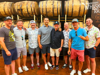 Group of eight men in casual summer outfits smiling and posing inside a Tennessee distillery warehouse in front of stacked oak whiskey barrels during a whiskey tour.