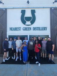 Twelve people posing for a group photo outside a distillery entrance under a large green logo on a white wooden facade, casual summer outfits and smiling faces, tour/tasting visit vibe.