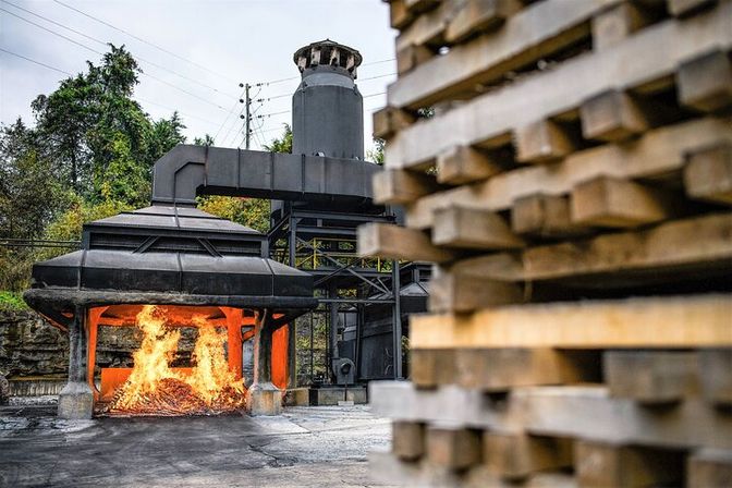 Open industrial kiln aglow with blazing wood fire, metal smokestack and ducting, with stacked lumber blurred in the foreground at an outdoor plant.