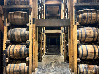 Towering wooden racks of stacked oak whiskey barrels in an aging warehouse, with a central concrete aisle receding into dim storage rows.