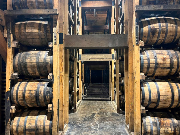 Towering wooden racks of stacked oak whiskey barrels in an aging warehouse, with a central concrete aisle receding into dim storage rows.