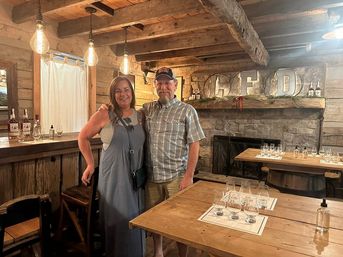 Smiling couple posing in a rustic wood-beam tasting room with Edison bulbs, a wooden bar, tables set with whiskey tasting glasses, and a stone fireplace.