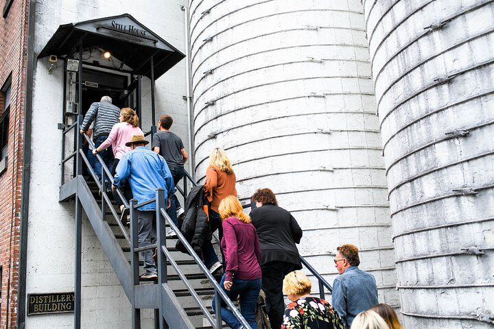 Tour group climbing an exterior metal staircase into an industrial distillery entrance flanked by large cylindrical concrete silos