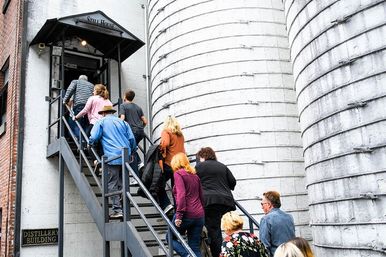 Tour group climbing an exterior metal staircase into an industrial distillery entrance flanked by large cylindrical concrete silos