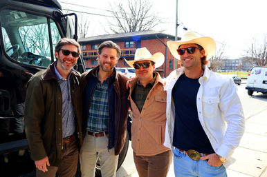 Four smiling men in casual jackets and sunglasses—two wearing cowboy hats—posing by a black tour van on a sunny urban street with brick apartment buildings and bare trees.