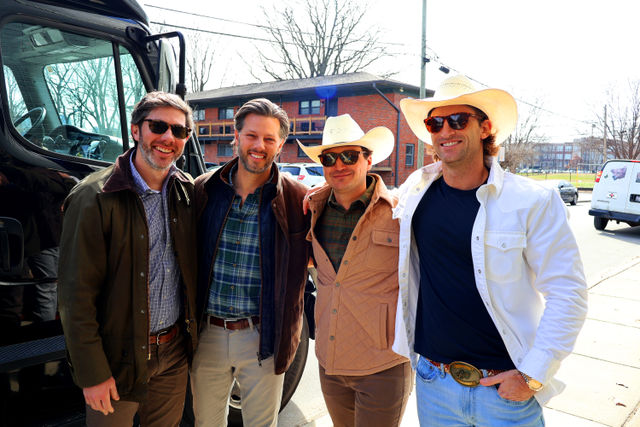 Four smiling men in casual jackets and sunglasses—two wearing cowboy hats—posing by a black tour van on a sunny urban street with brick apartment buildings and bare trees.