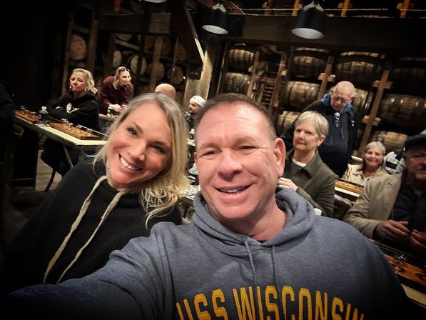 Smiling couple taking a selfie in a cozy whiskey distillery barrel room, stacked oak barrels behind them and other visitors seated at long tables sampling tastings.