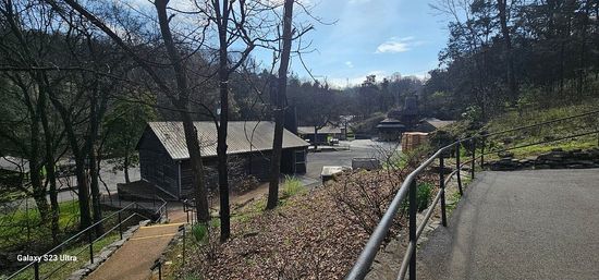 Hilly park scene with paved walkway and metal railing leading past stone steps to a rustic wooden barn surrounded by leafless trees under a bright blue sky.