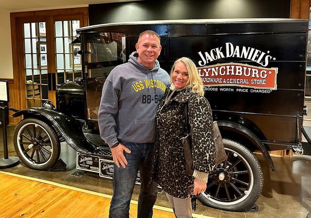 Smiling couple posing indoors beside a black vintage delivery truck with a large logo panel in a museum-style exhibit