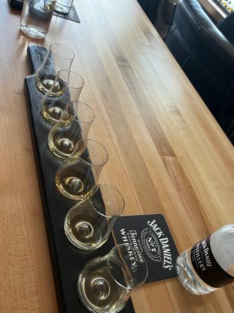 Whiskey tasting flight of six tulip-shaped glasses with small amber pours on a black wooden paddle, arranged along a light wood bar with a water bottle and coaster nearby.