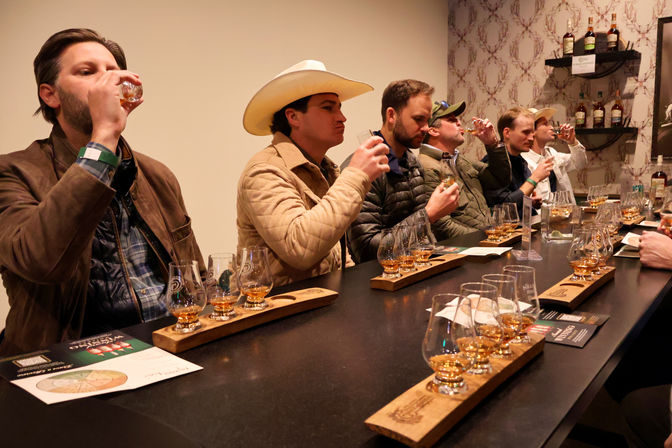 Whiskey tasting at a distillery-style bar with a row of people sampling flights on wooden trays — multiple tasting glasses, bottles on shelves, and one guest in a cowboy hat.