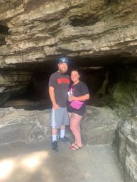 Couple smiling and posing inside a shallow limestone cave entrance with layered rock ledges, mossy walls and casual hiking gear.