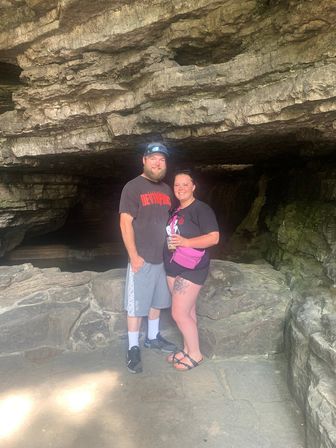 Couple smiling and posing inside a shallow limestone cave entrance with layered rock ledges, mossy walls and casual hiking gear.