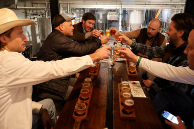 Group of six people clinking small beer tasting glasses over wooden flight boards in a craft brewery tasting room, stainless steel fermenters visible in the background.