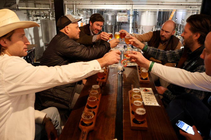 Group of six people clinking small beer tasting glasses over wooden flight boards in a craft brewery tasting room, stainless steel fermenters visible in the background.