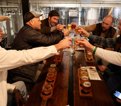 Group of six people clinking small beer tasting glasses over wooden flight boards in a craft brewery tasting room, stainless steel fermenters visible in the background.
