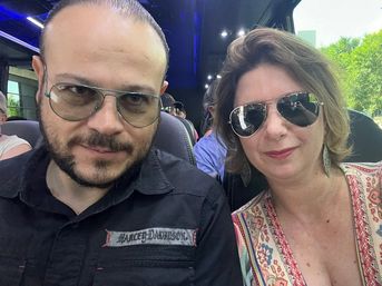 Two adults wearing sunglasses pose for a selfie on a tour bus, seated among other passengers with bus seats and green trees visible through the window.